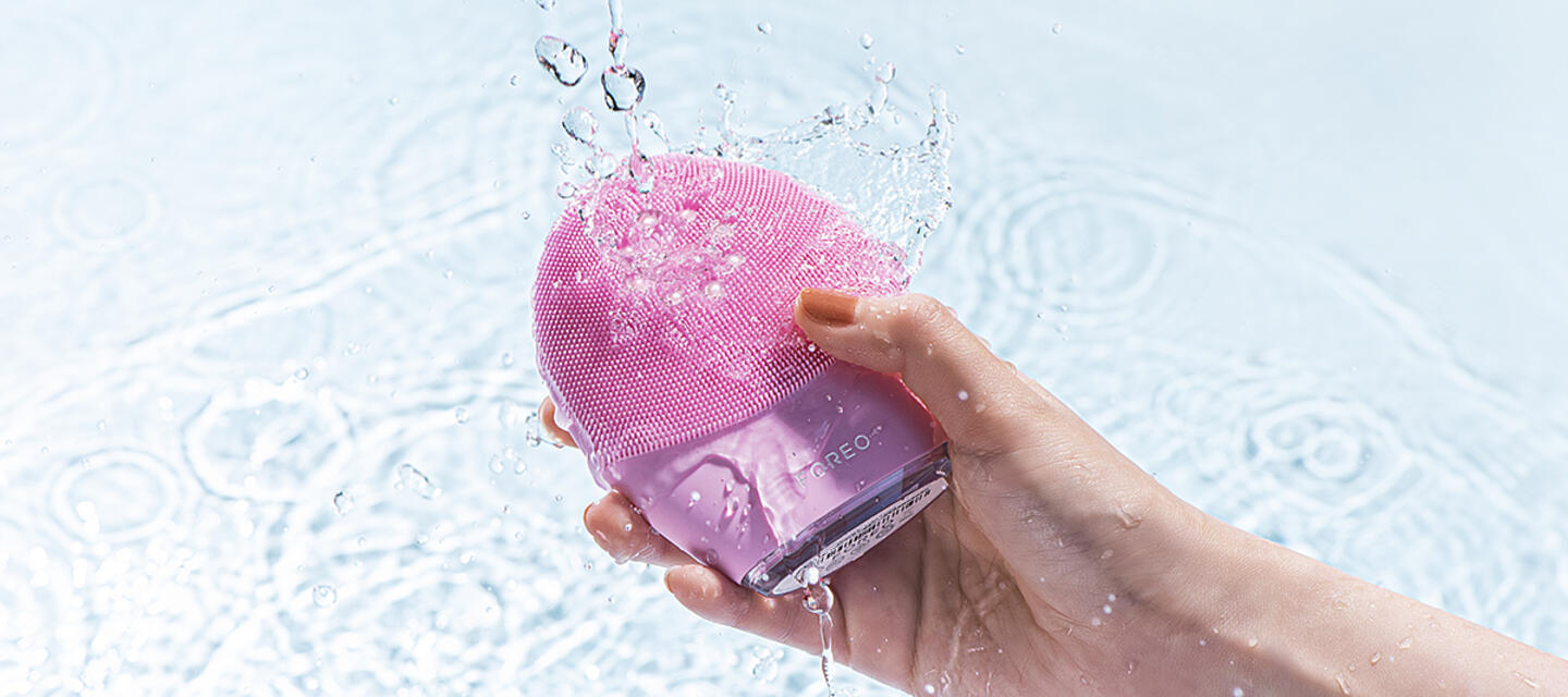 Hand holding FOREO LUNA 4 plus pink facial cleansing brush, surrounded by water